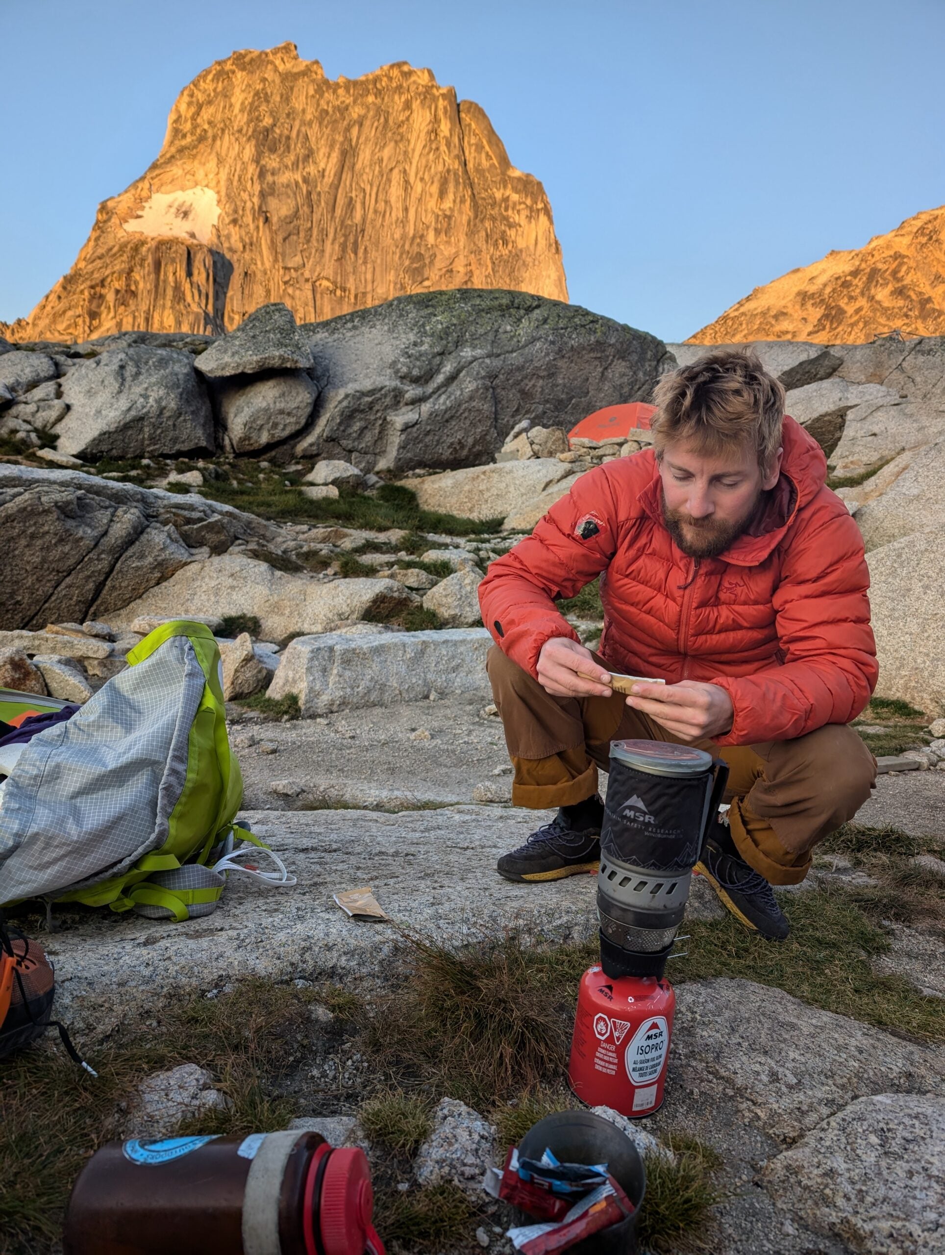Climber boils water with small camping stove at sunrise in front of a tall mountain.